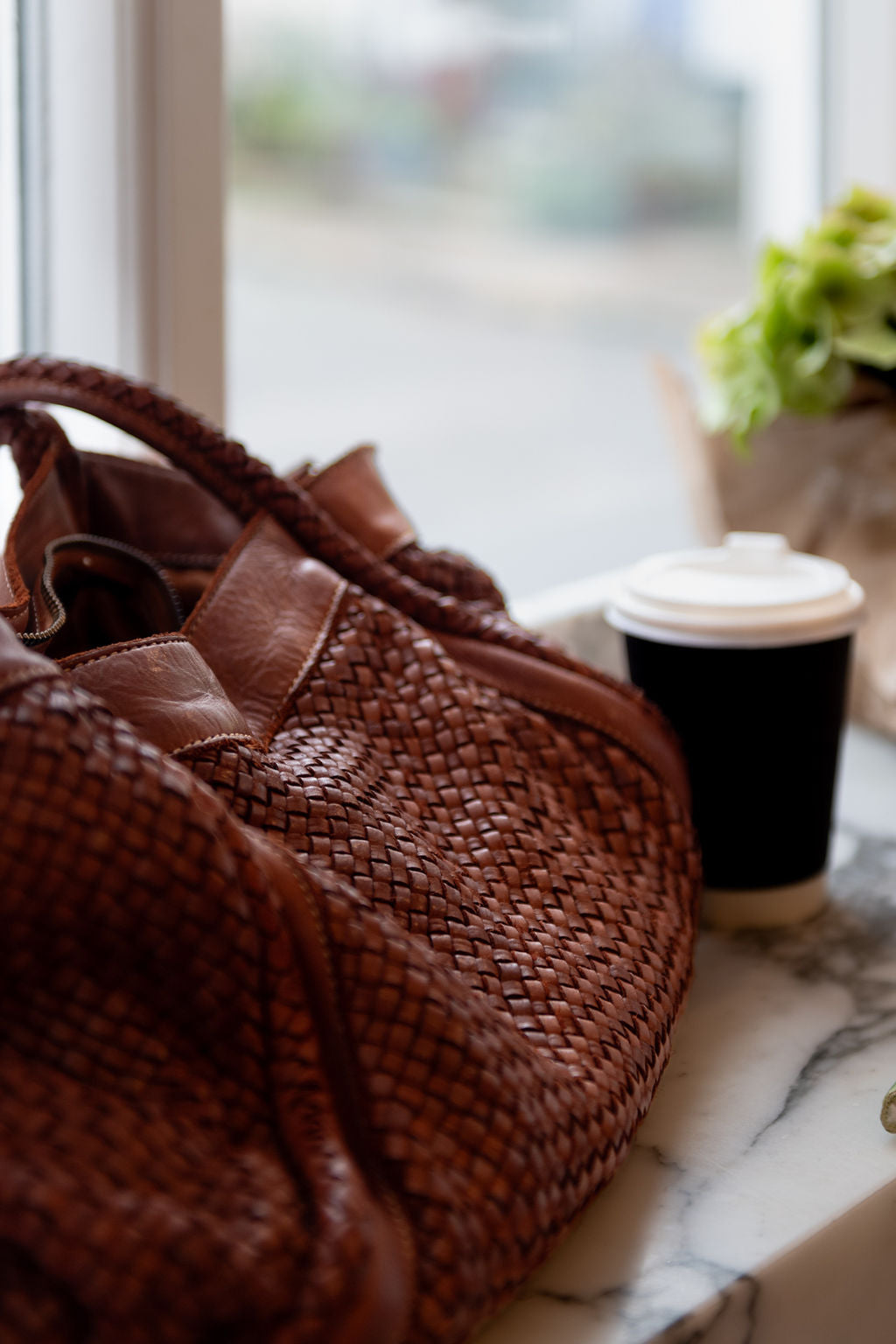Tan woven Italian leather handbag on a marble surface with a blurred window and plant in the background