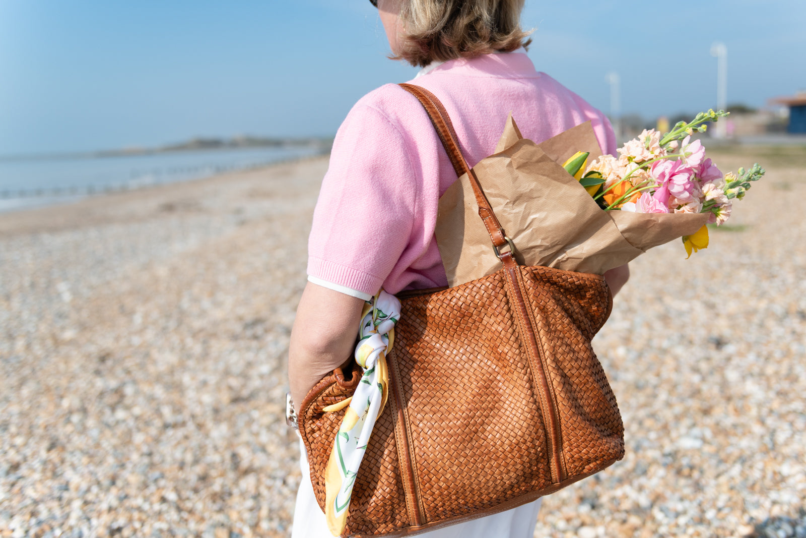 Person holding a woven brown bag and flowers on a beach