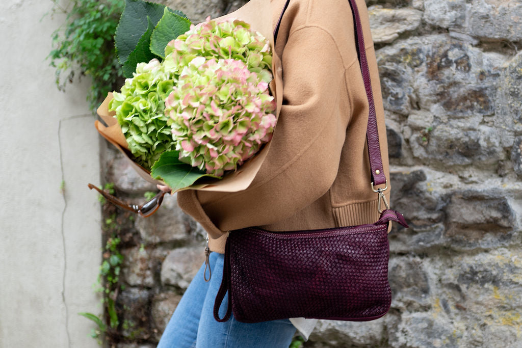 Person holding a bouquet of flowers with a purple handbag against a stone wall.