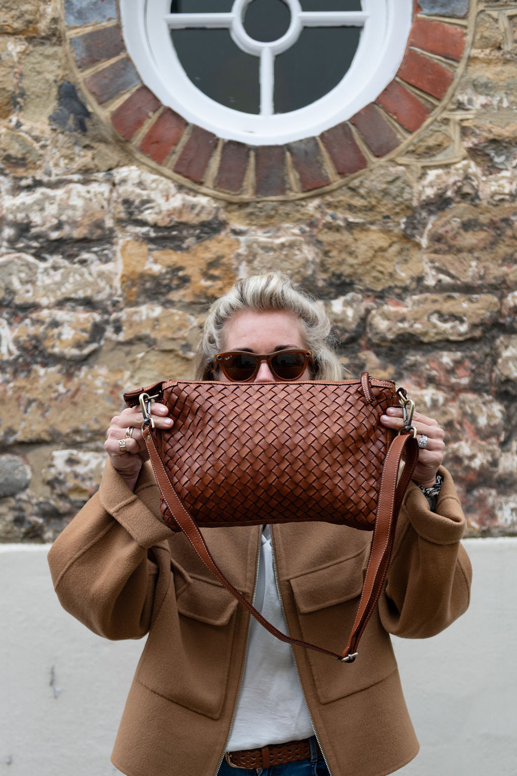 Person holding a tan colour hand woven Italian leather handbag in front of a stone wall. 